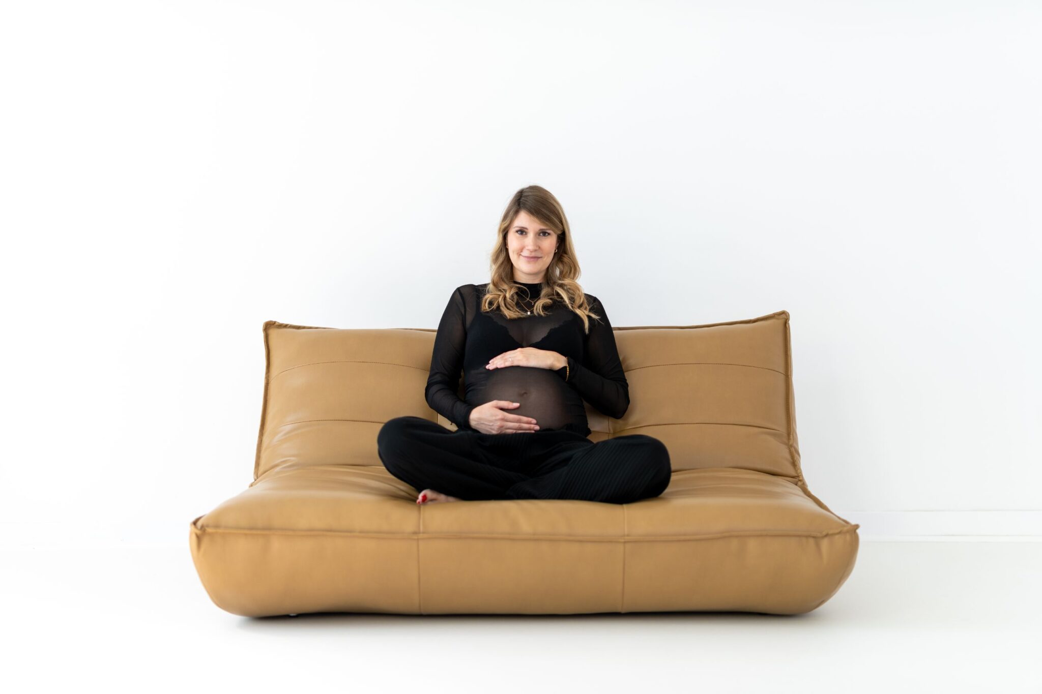 Schwangere Frau sitzt mit gekreuzten Beinen auf einem beigen Sofa und hält ihren Babybauch, weißer Hintergrund.