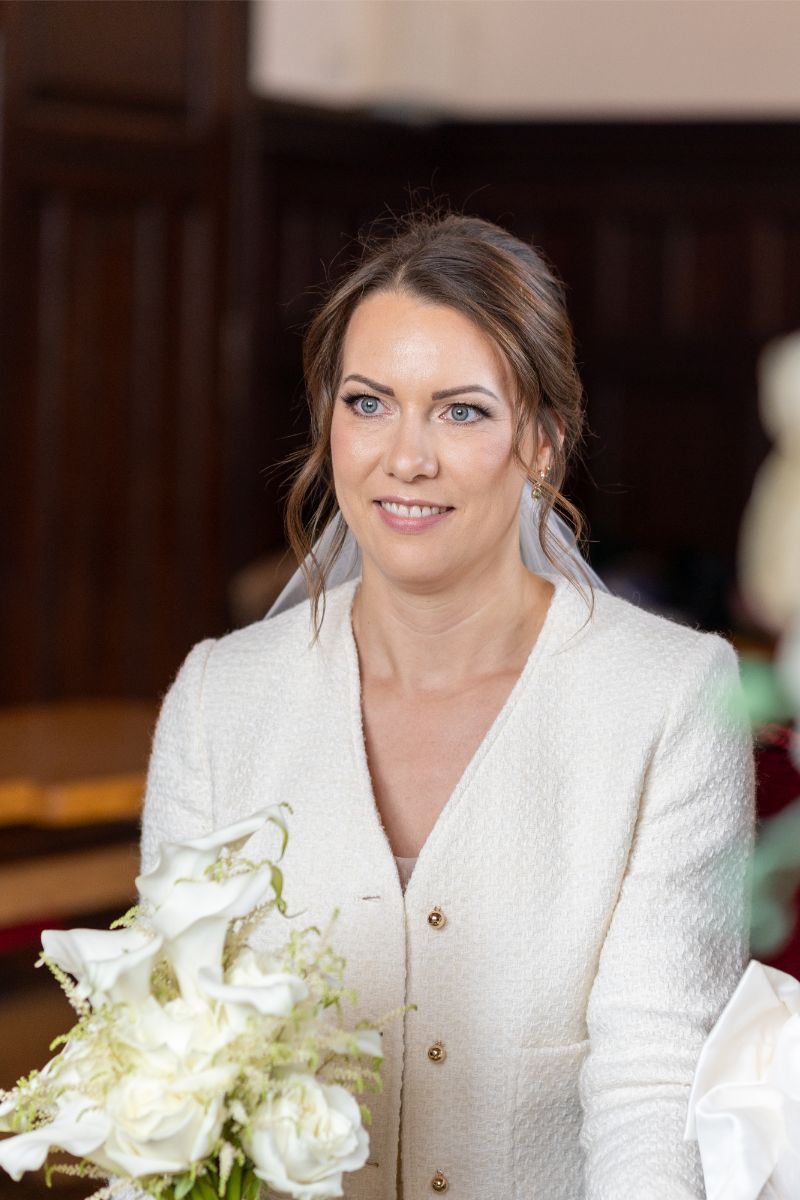 Frau mit braunen Haaren im weißen Blazer hält einen Blumenstrauß und lächelt in einem festlich geschmückten Raum.