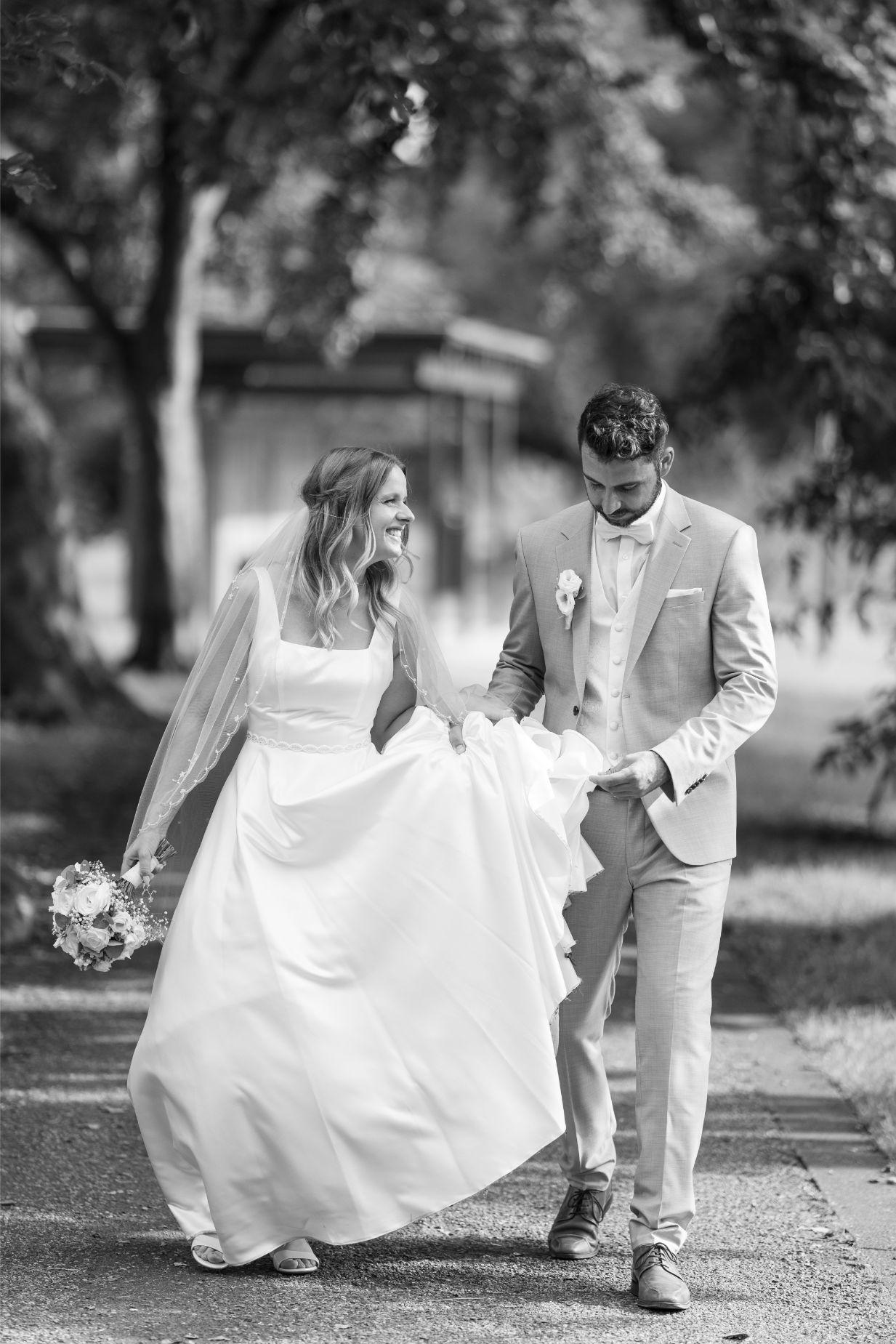 Ein Brautpaar spaziert draußen. Die Braut trägt ein weißes Kleid und hält Blumen, der Bräutigam trägt einen hellen Anzug. Schwarz-weißes Foto.