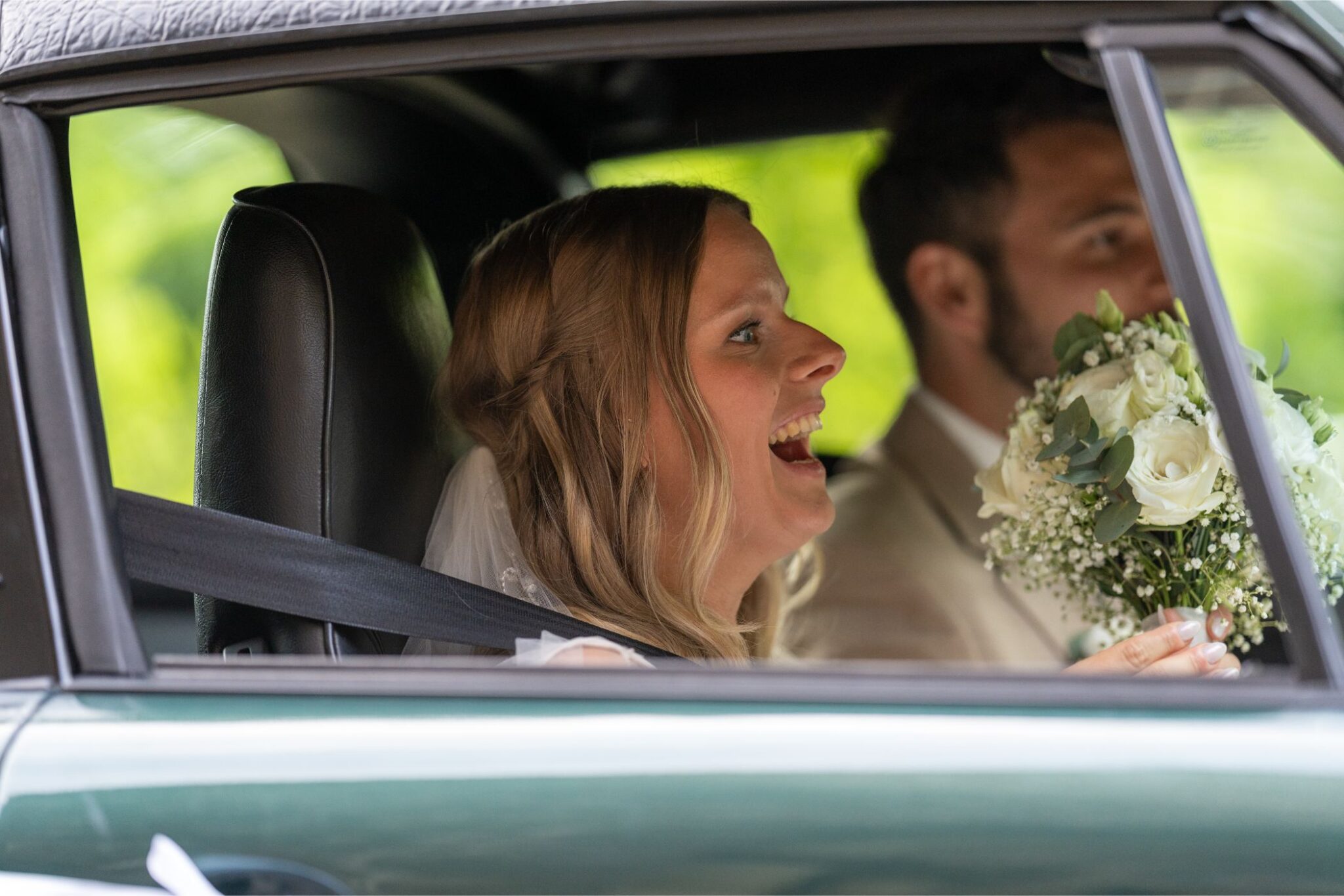 Eine Braut und ein Bräutigam sitzen lächelnd im Auto, die Braut hält einen Blumenstrauß und trägt einen Schleier.