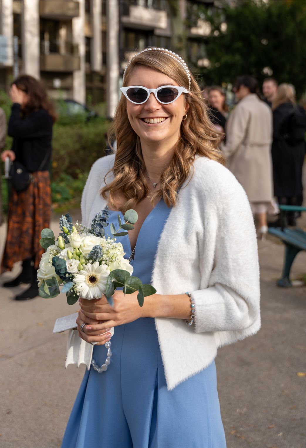 Frau mit weißen Sonnenbrillen, blauem Kleid und weißer Strickjacke hält einen Blumenstrauß, lächelt und steht im Freien mit anderen Personen im Hintergrund.
