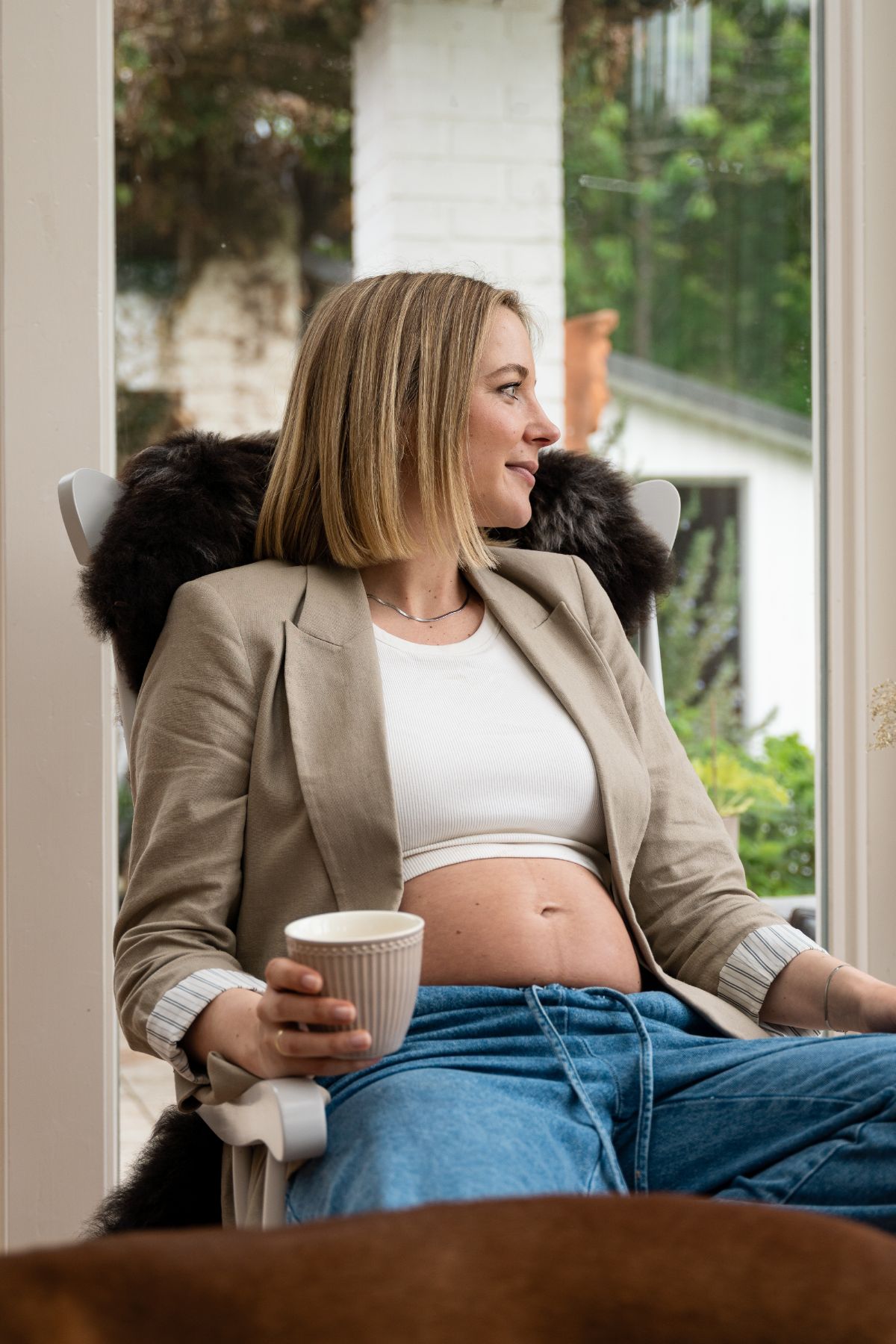 Schwangere Frau sitzt entspannt auf einem Stuhl, hält eine Tasse in der Hand und blickt aus dem Fenster.