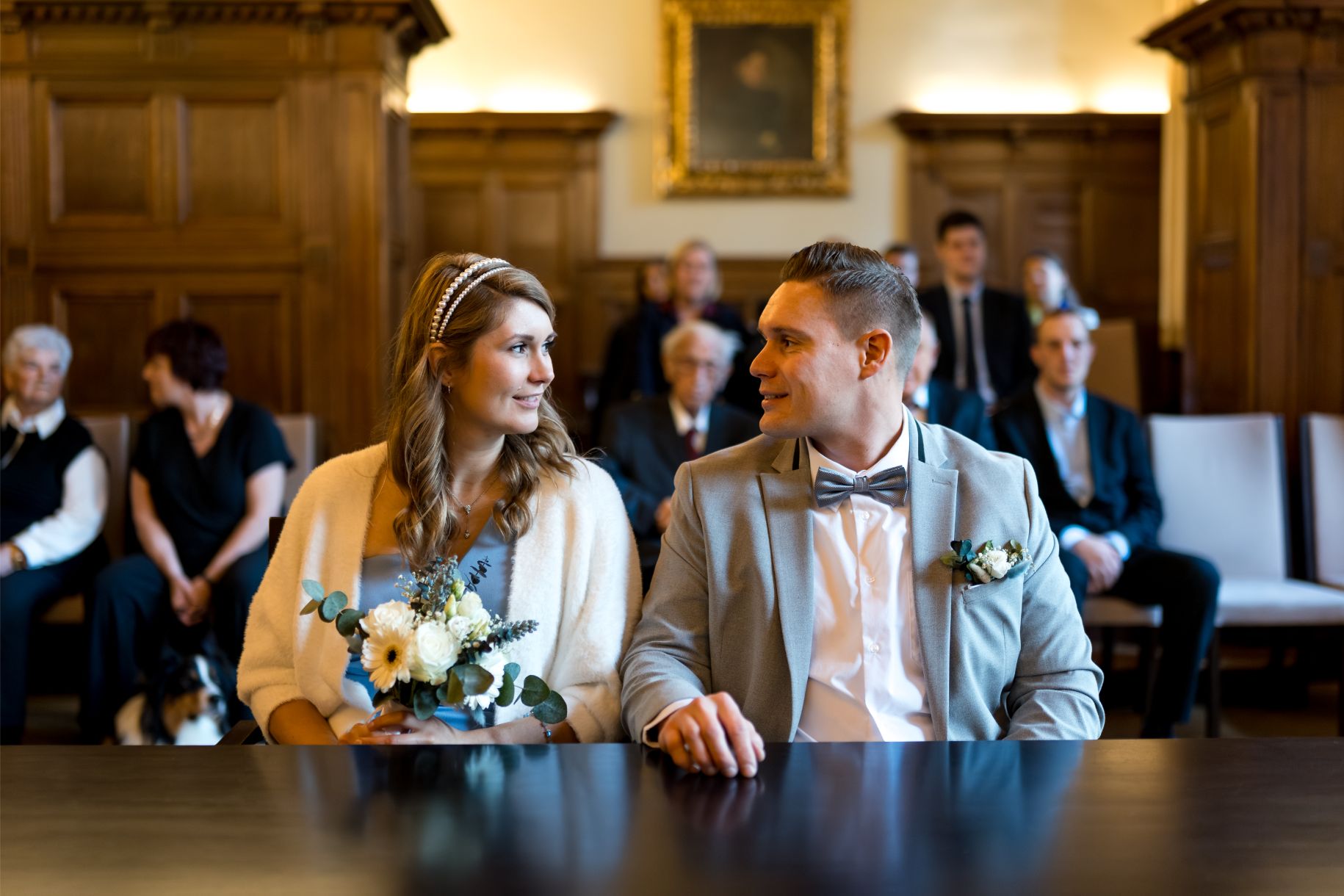 Ein Brautpaar sitzt nebeneinander an einem Tisch im Standesamt, beide schauen sich an. Gäste sitzen im Hintergrund.
