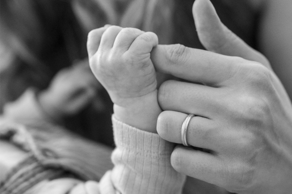 Ein Baby hält den Finger eines Erwachsenen fest. Die Hand des Erwachsenen trägt einen Ring. Schwarz-weiß Fotografie.