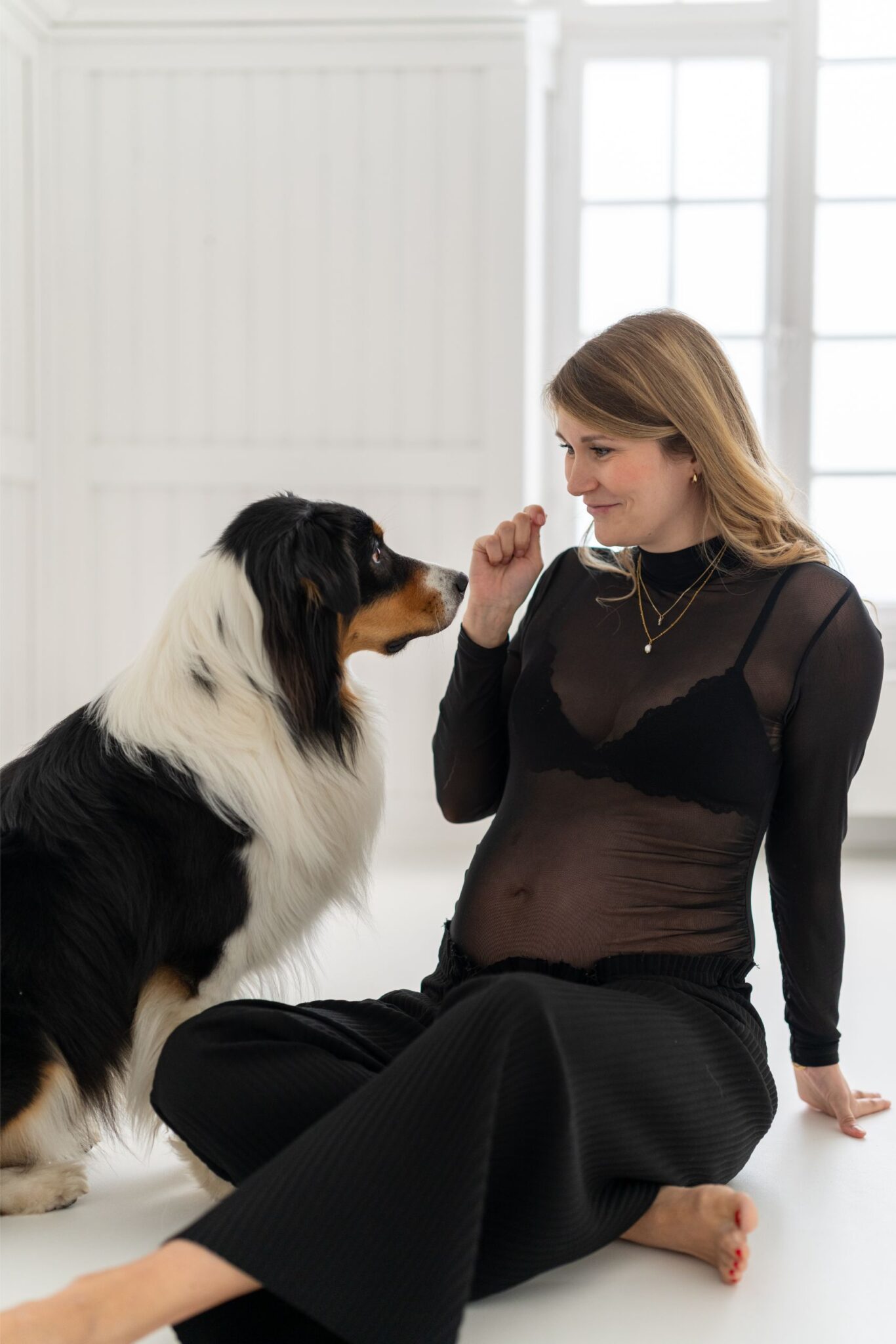 Eine Frau im schwarzen Outfit sitzt auf dem Boden und schaut lächelnd einen sitzenden Hund an, der sie aufmerksam beobachtet, in einem hellen Raum.
