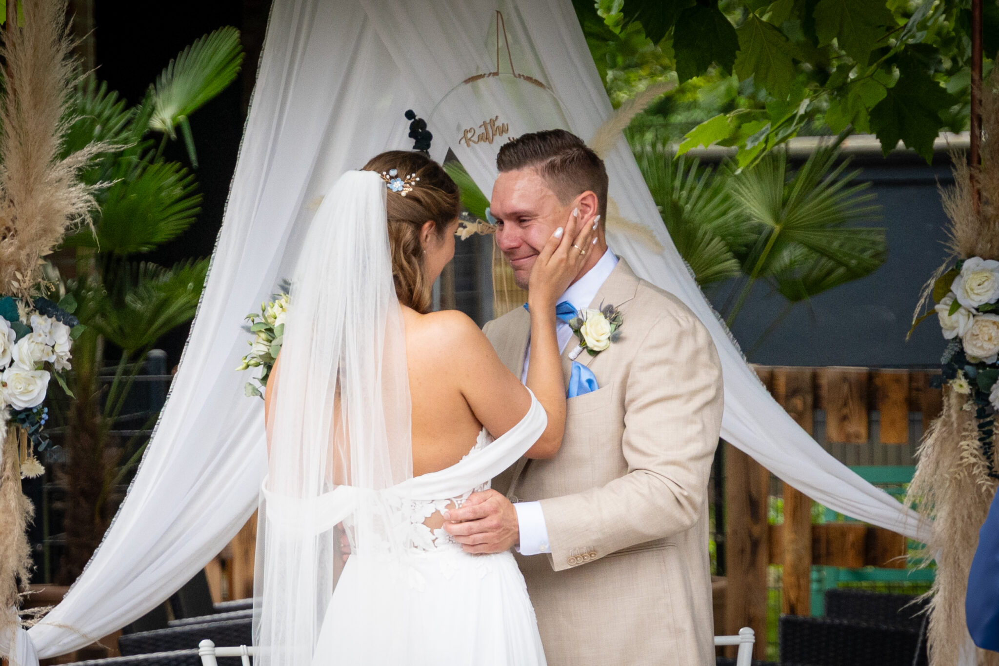 Ein Brautpaar steht sich lächelnd gegenüber, die Braut berührt zärtlich das Gesicht des Bräutigams, im Hintergrund weiße Hochzeitsdekoration und grüne Pflanzen.